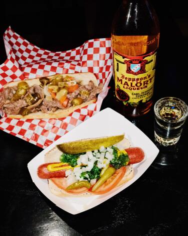 An Italian beef sandwich with a Chicago dog and a bottle and shot of Jeppson's Mal?rt on a black table at Tiny's Hi-Dive.