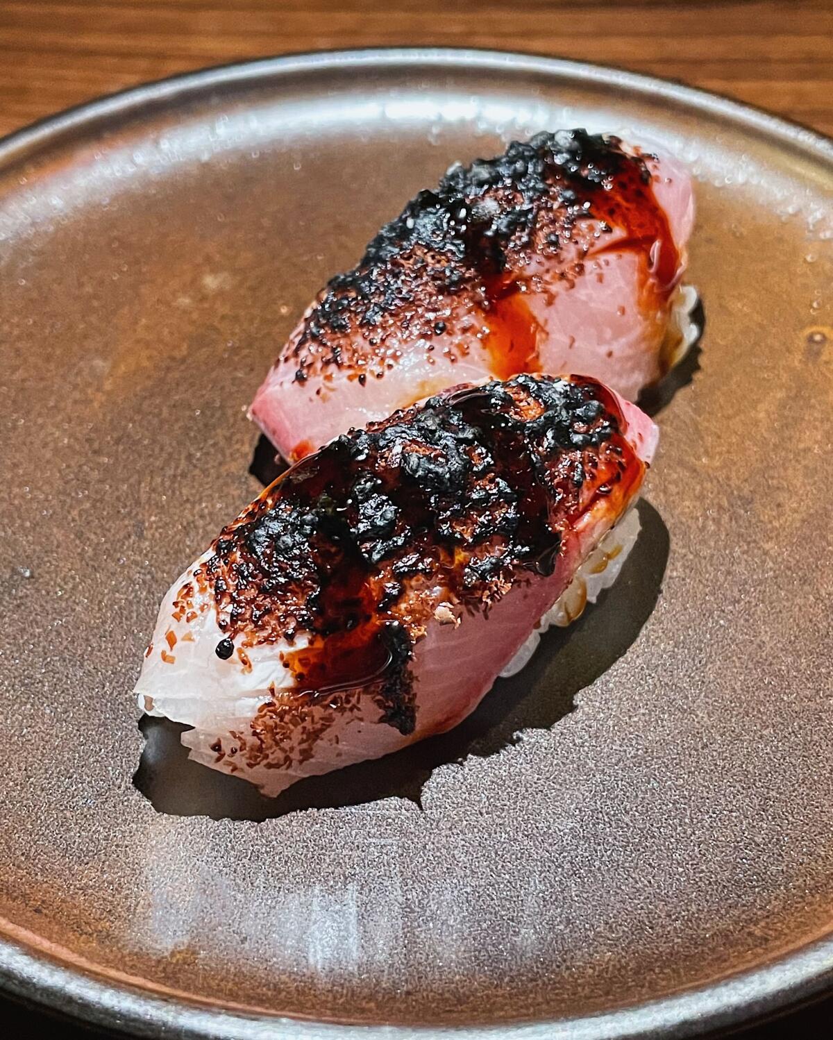 Two pieces of hamachi nigiri topped with Oaxacan chocolate on a brown ceramic plate