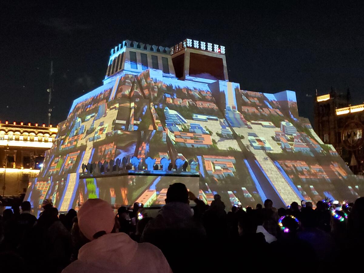 A brightly lighted work of art shown at night is surrounded by a crowd of people.