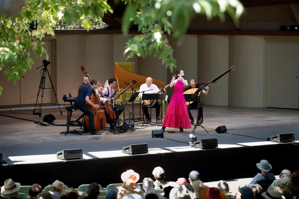 A group of seated musicians performs on a stage while a standing woman plays a flute.