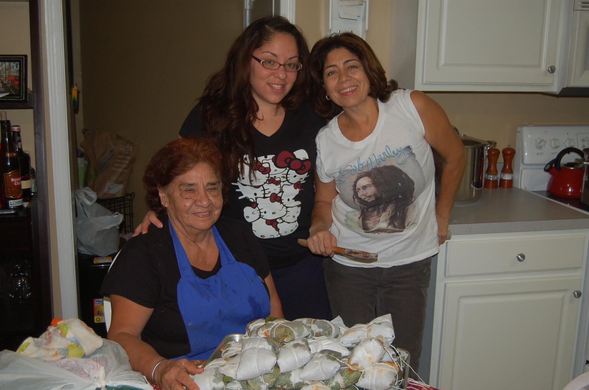 Three women, two standing and one seated at a table piled high with tamales