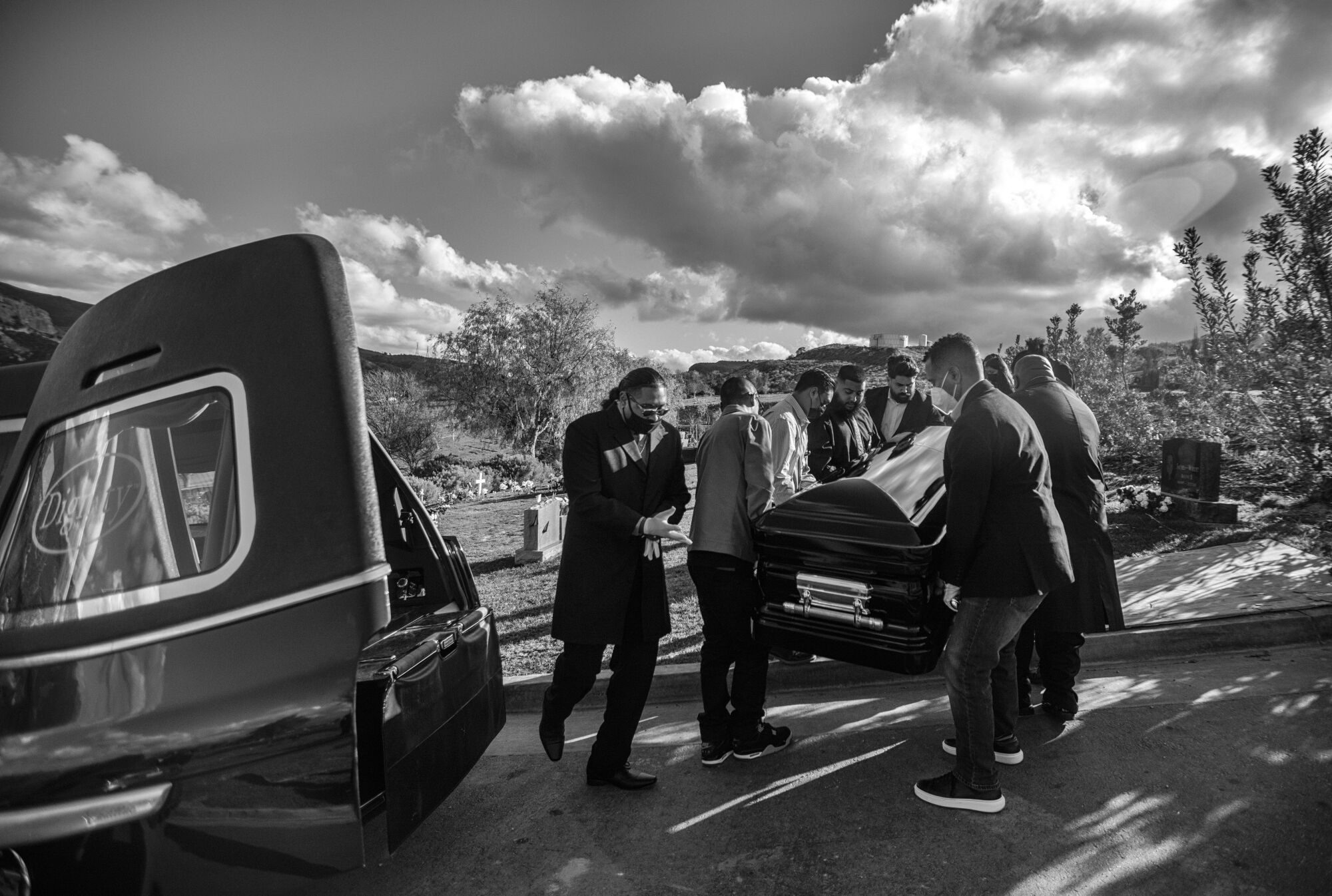 Pallbearers carry a casket toward a hearse under a cloudy sky