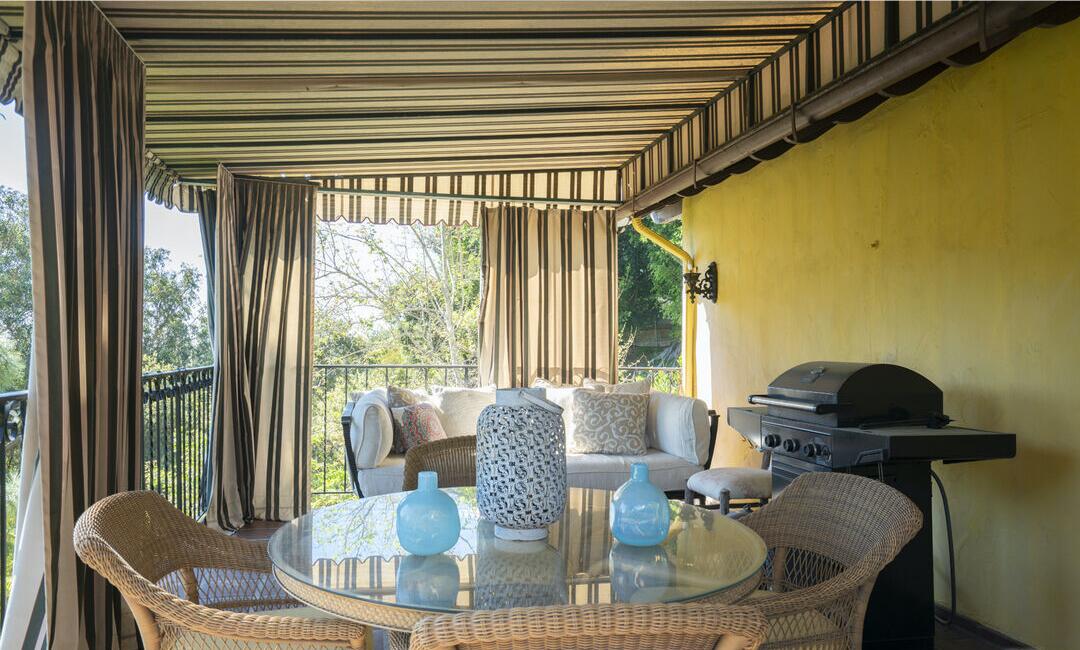 The terrace has striped drapes and two tables with wicker seats overlooking greenery.