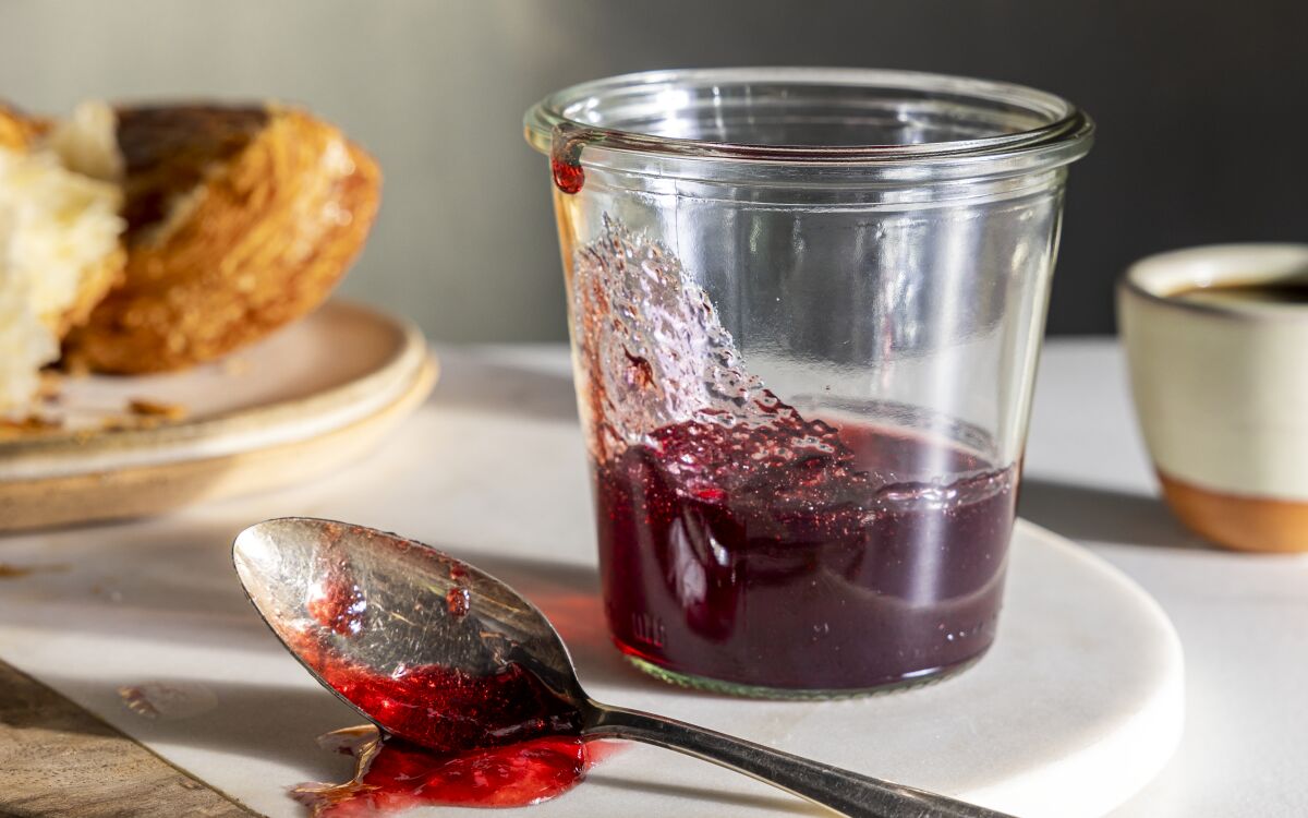 A spoon with jelly on it rests next to a clear glass jar partly filled with jelly.
