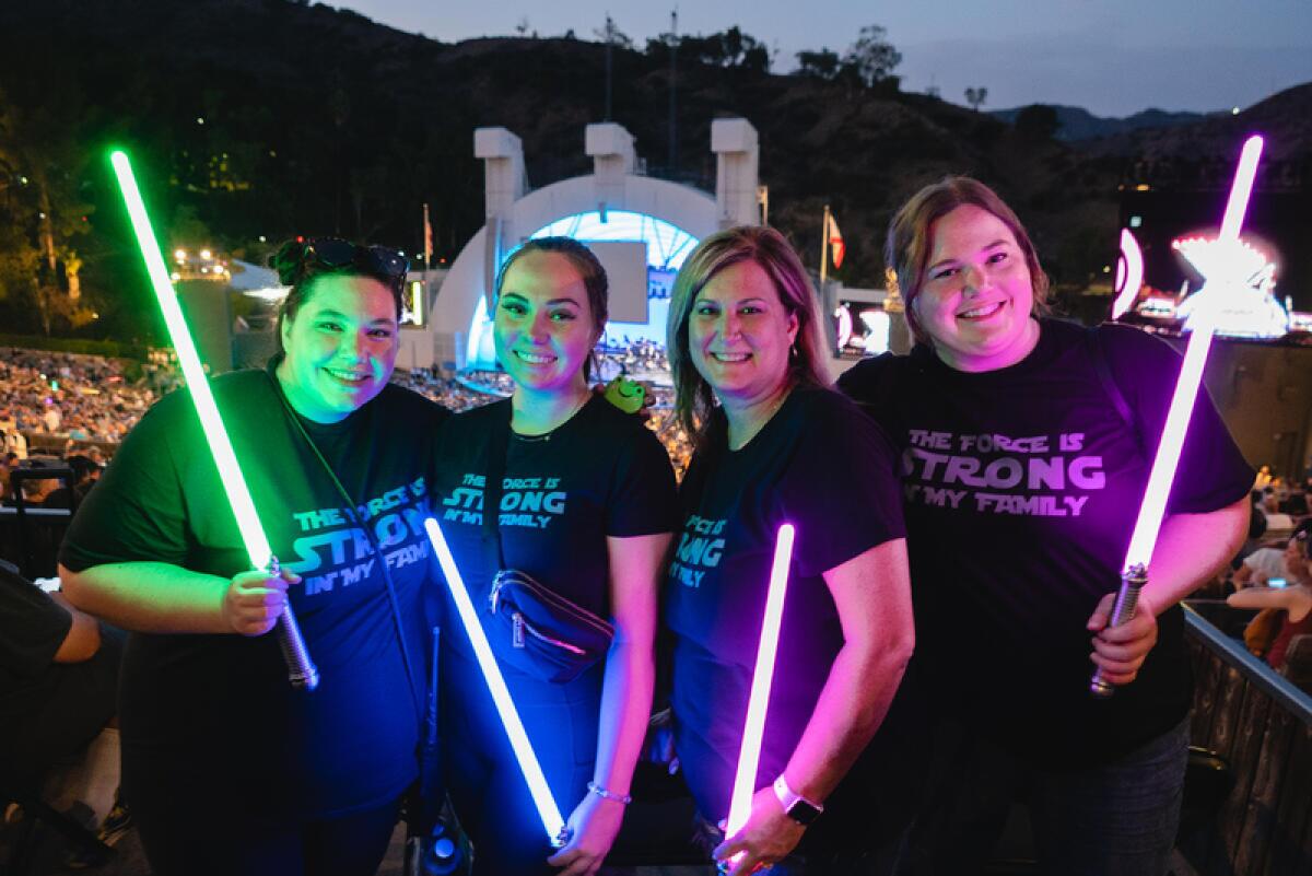 Four people smiling and holding lightsabers in front of the Hollywood Bowl stage.