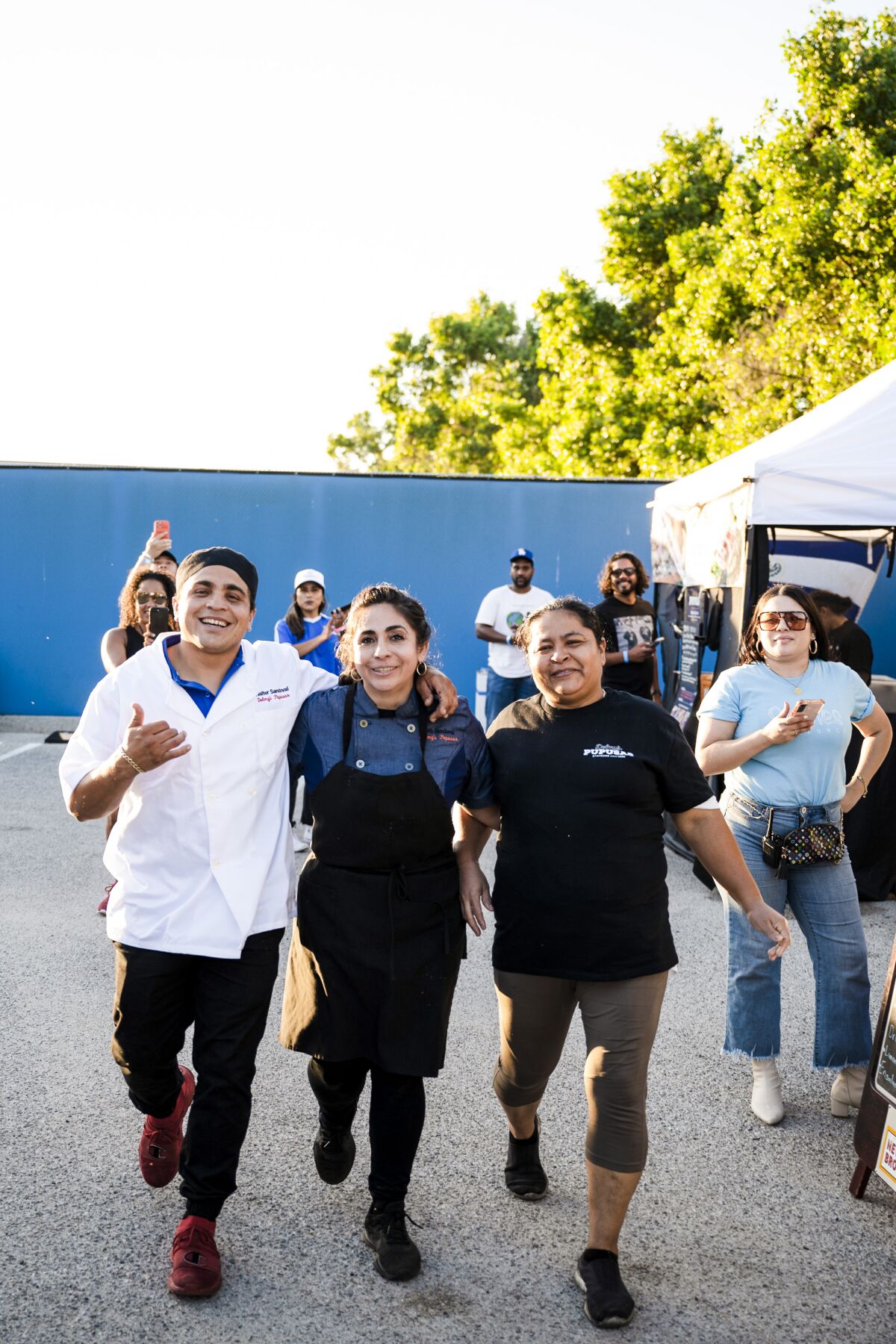 Three people, one in an apron and another in a chef's jacket, smile as they walk with arms linked.