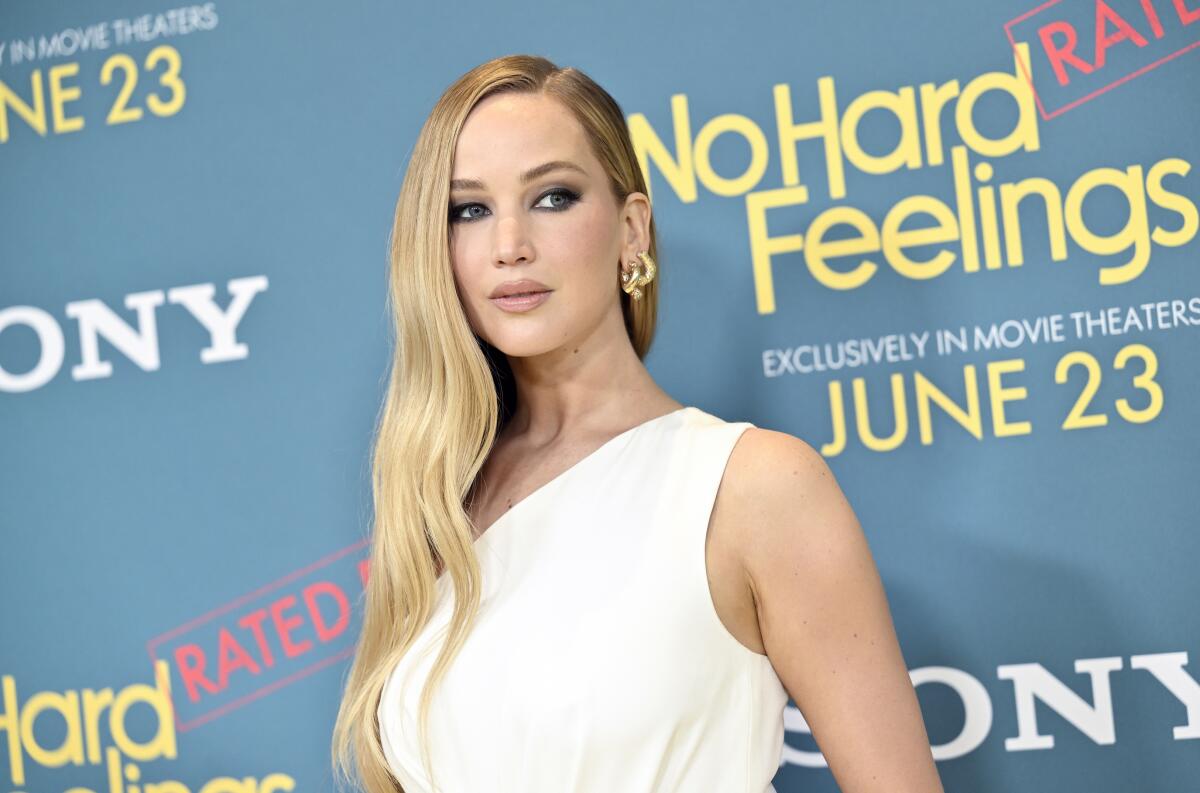 Jennifer Lawrence has long blond hair and wears a white dress posing in front of a backdrop that says "No Hard Feelings."