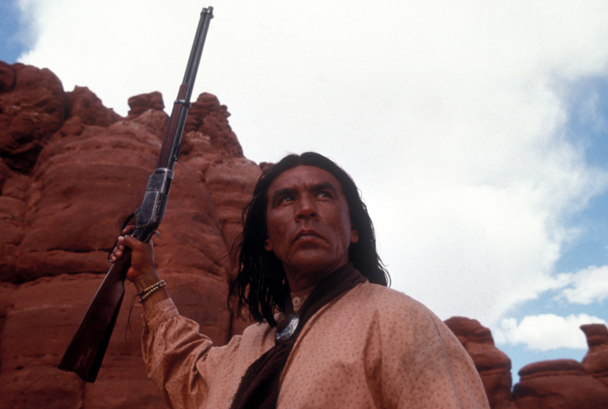 A Native American hoists a rifle in a canyon.