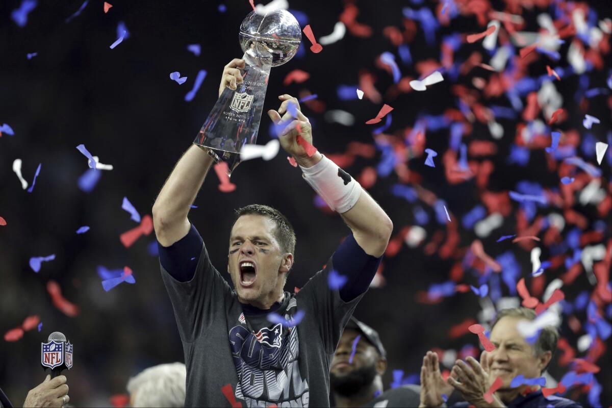 Tom Brady raises a silver trophy shaped like a football on a pedestal as confetti falls