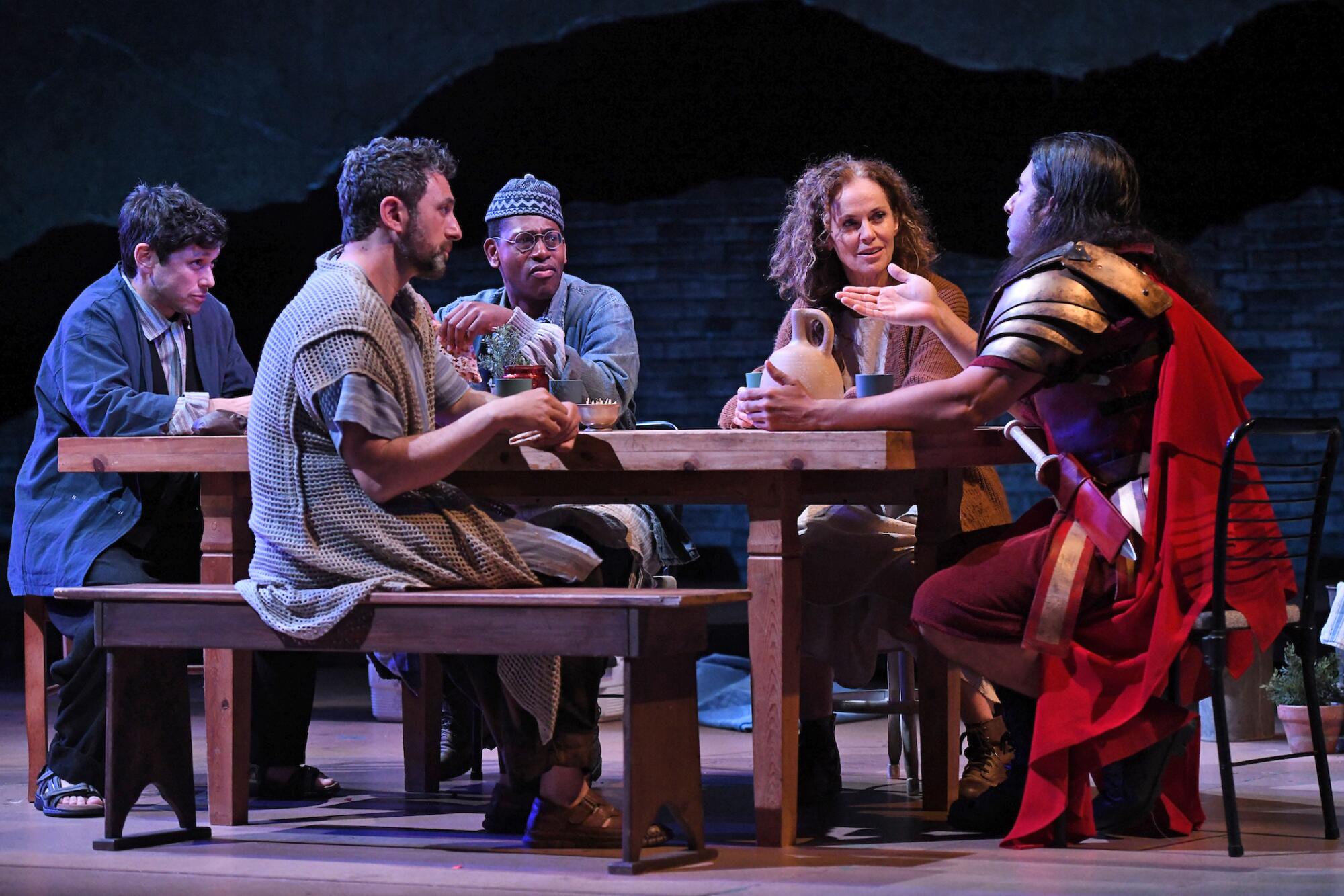 A person dressed as a Roman centurion talks to four people sitting around a table onstage