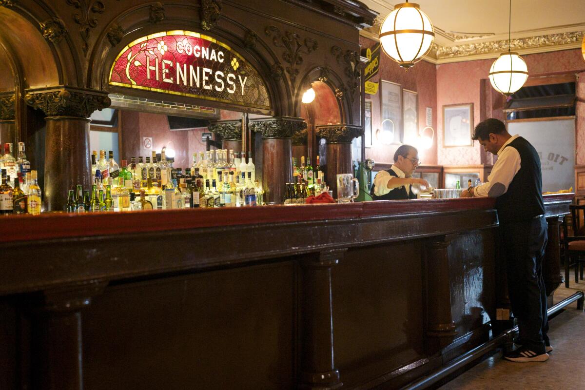 Two men stand by the bar. Bottles sit below a sign reading "Cognac Hennessy."
