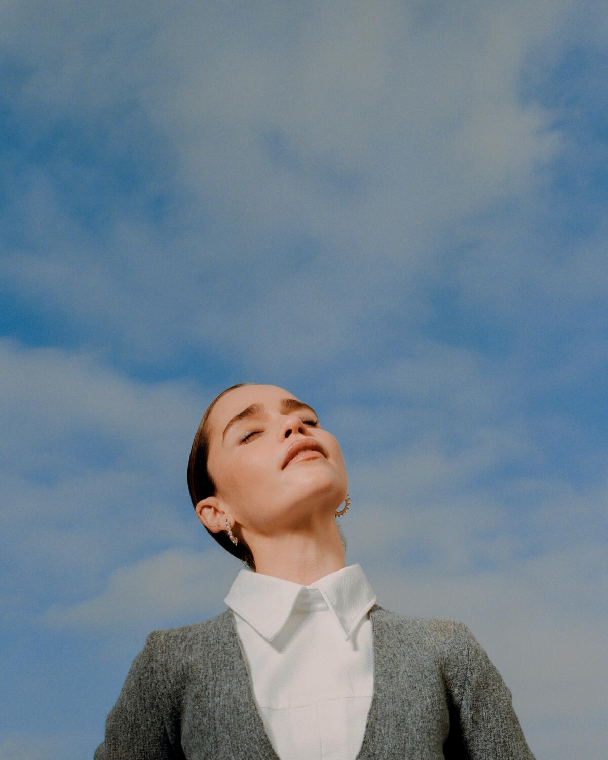 Emilia Clarke looks upward toward a blue cloudy sky.