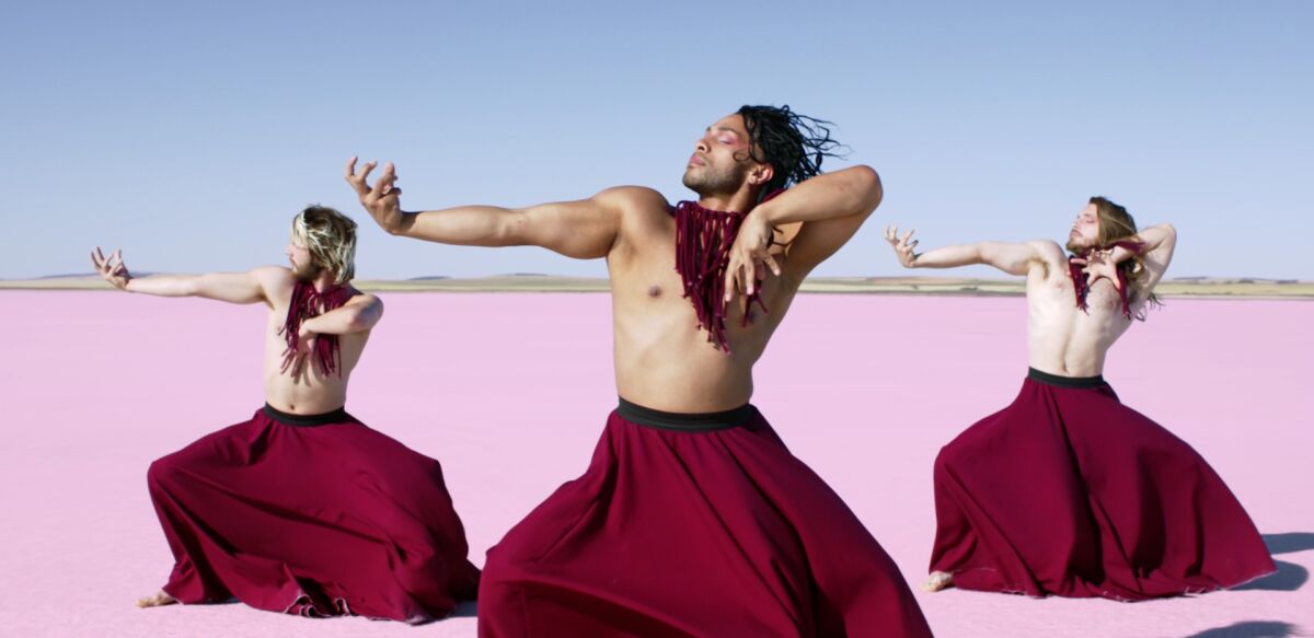 Three men in broad red skirts dance against a pink landscape