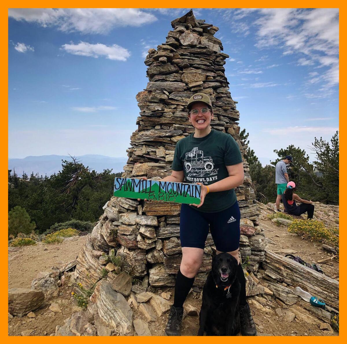 A person holds a sign that says Sawmill Mountain in front of a pile of stacked rocks and next to a dog.