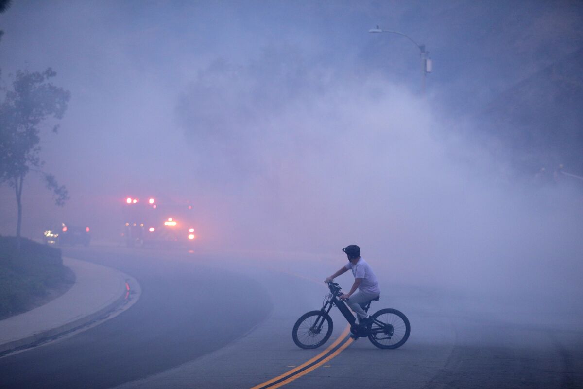 A bicyclist pauses in a roadway, looking at a fire truck whose lights are visible in the smoky gloom.