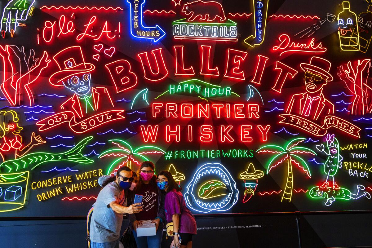 A group take a selfie in front of a neon sign for Bulleit Frontier Whiskey inside the Grand Central Market.