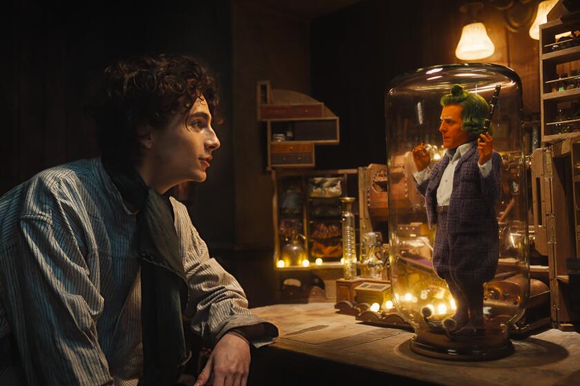 Timothée Chalamet as Willy Wonka looks into a glass jar containing an Oompa Loompa played by Hugh Grant