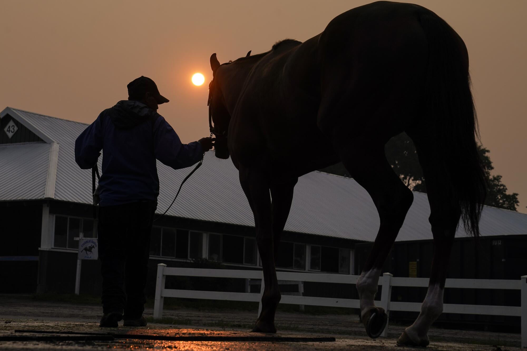 A silhouetted handler leads a horse back into the stables as the sun is obscured by haze.