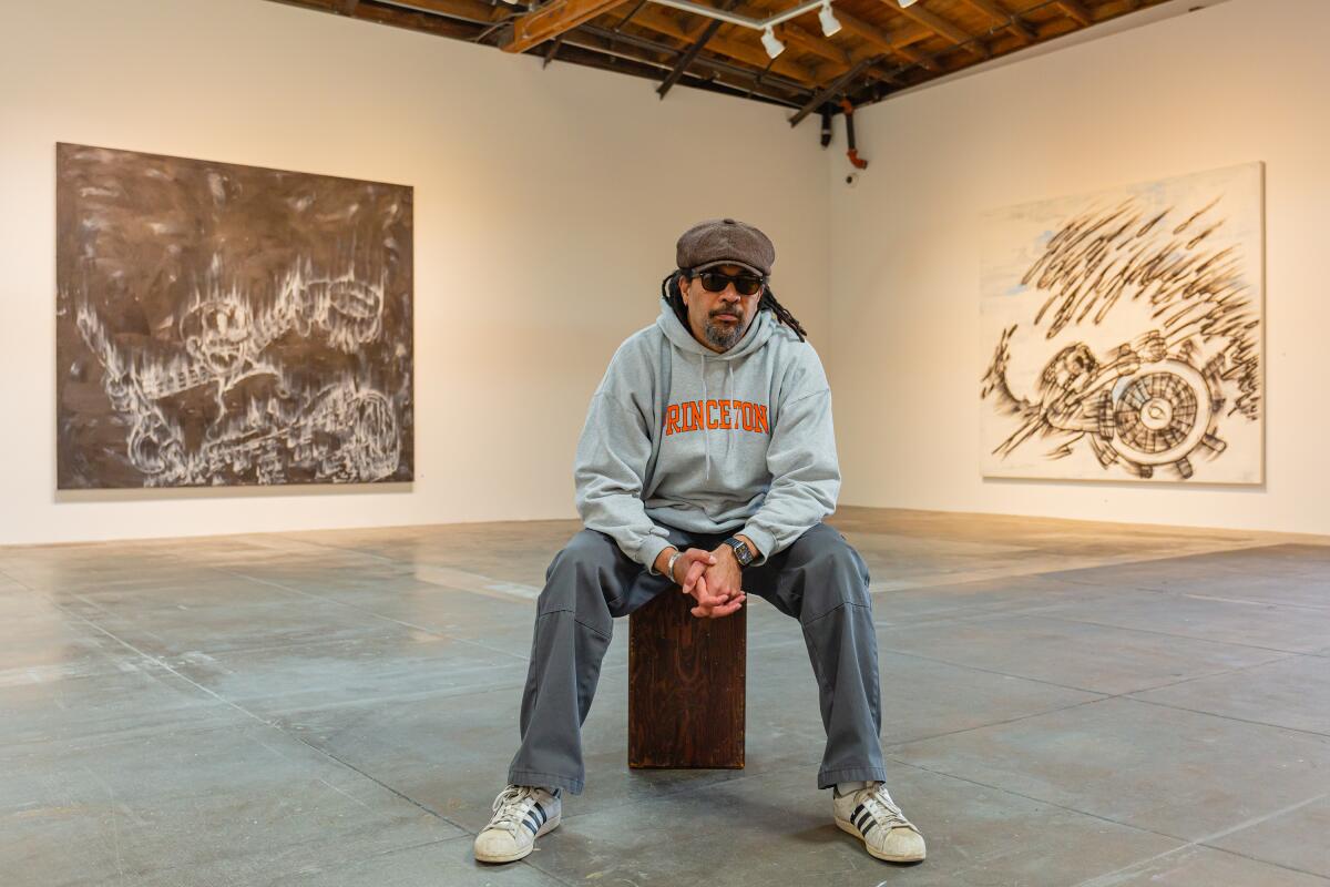 A man in a Princeton hoodie and newsboy cap sits among paintings.