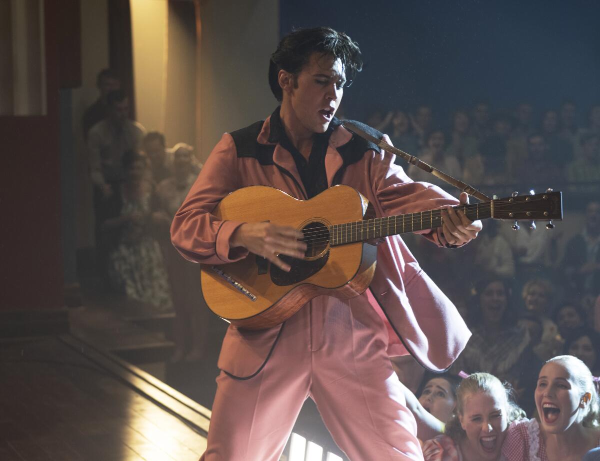 A male actor wears a pink suit and plays a guitar on stage in a scene from a movie.