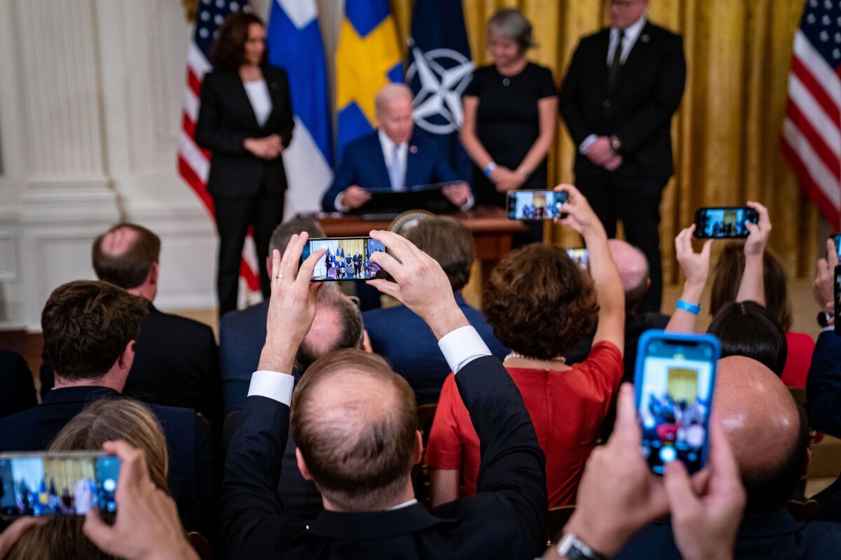 People hold up cellphones facing a person sitting at a desk.