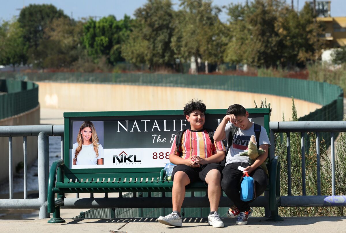 Two boys sit on a green bus bench over a river channel in the hot afternoon sun.