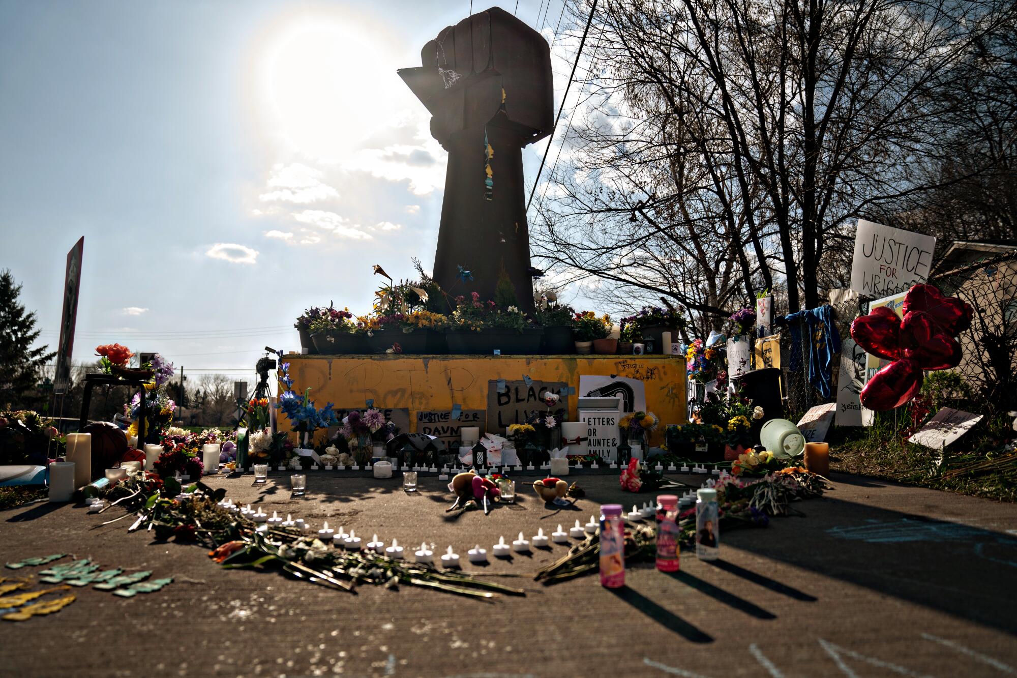 Candles, flowers, signs and other items sit on the ground before a statue of a raised fist