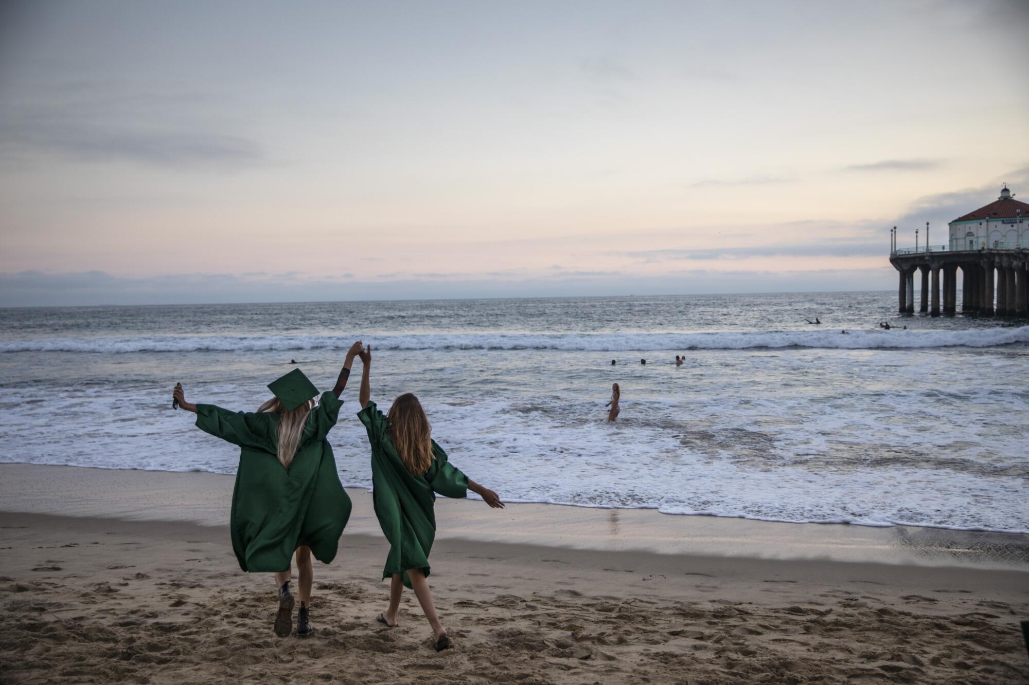 Two girls in graduation gowns walk on the sand near a pier, their clasped hands upraised.