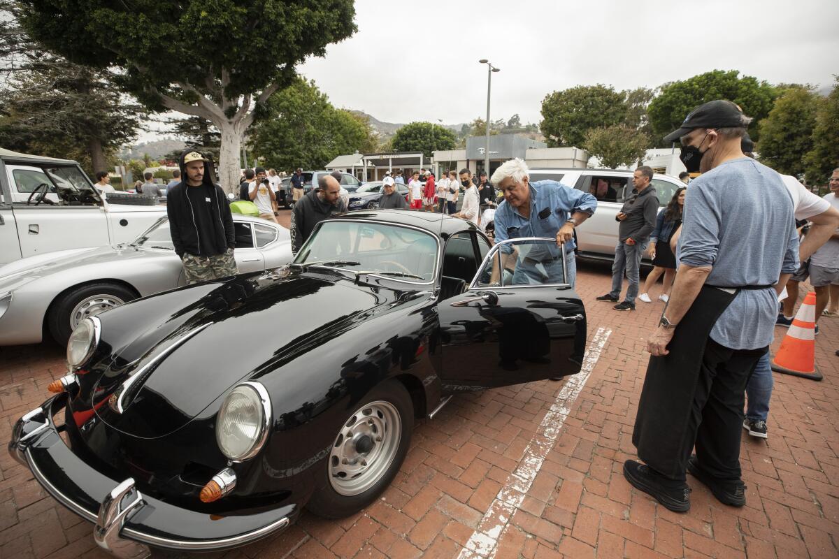 People stand around a black vintage car as a white-haired driver opens the driver-side door.