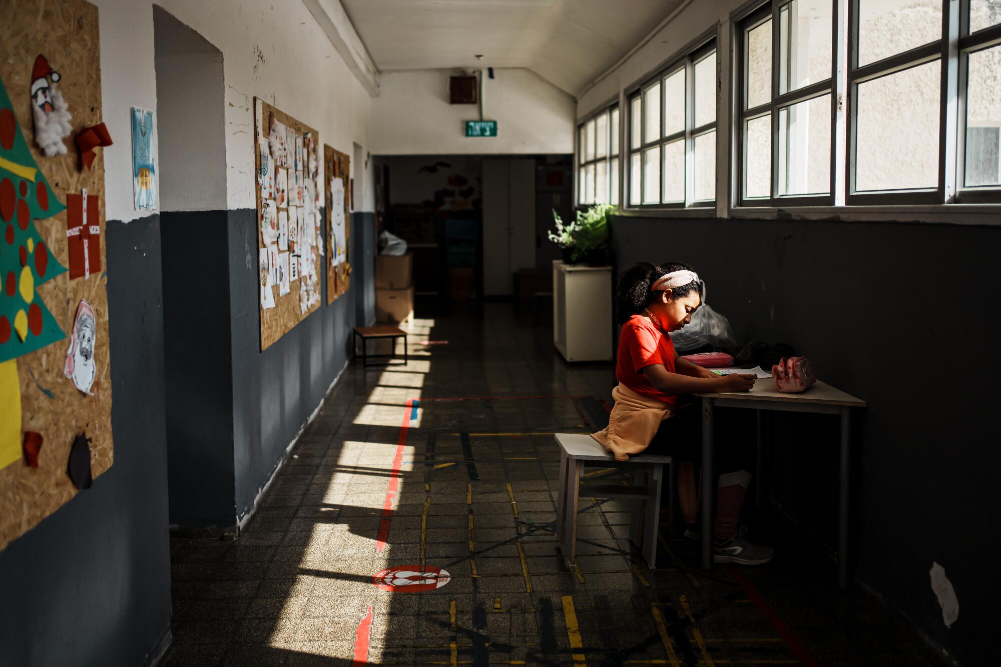 A young girl seated at a desk in a hallway under a row of windows 