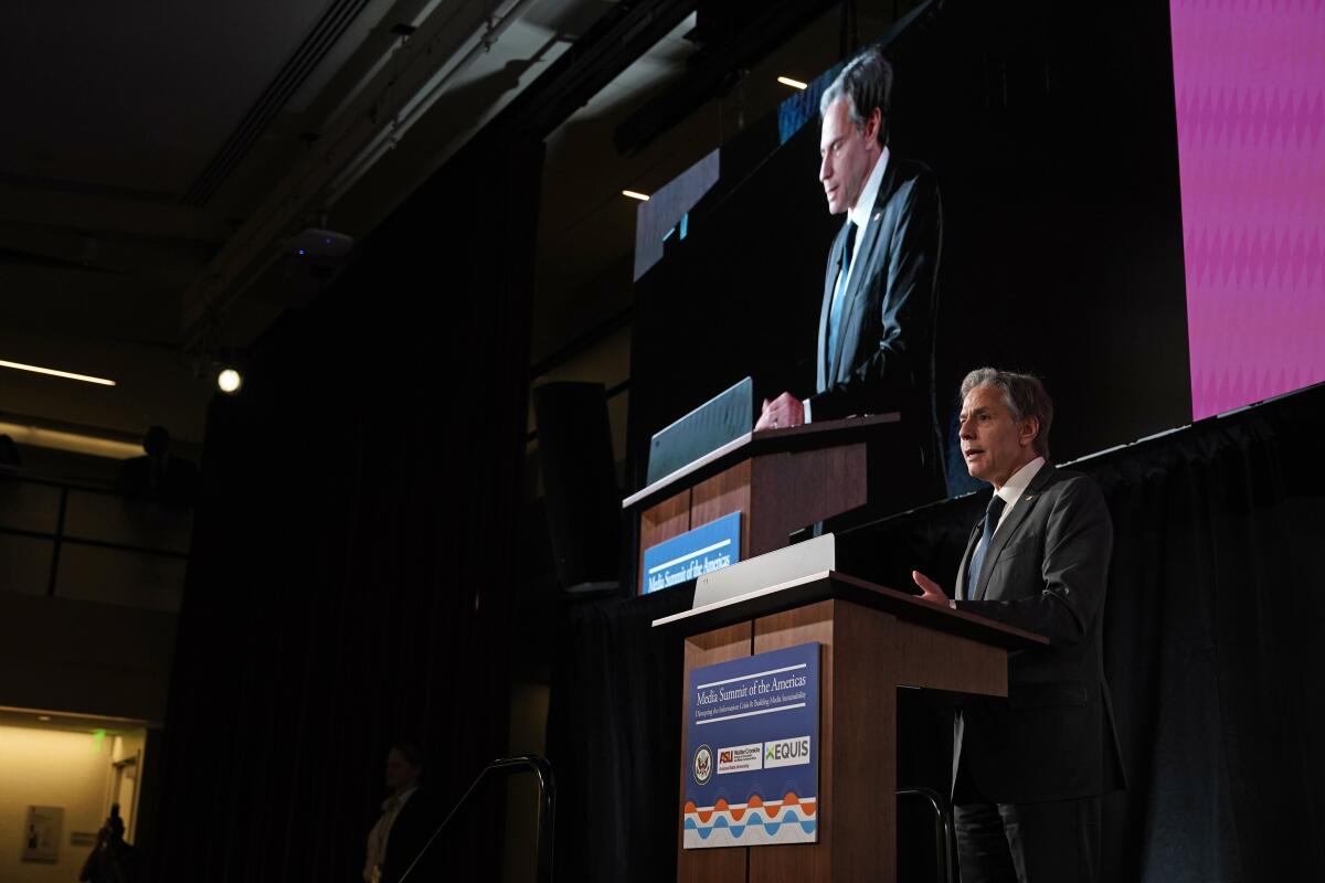 Secretary of State Anthony Blinken speaks next to a giant screen showing his image