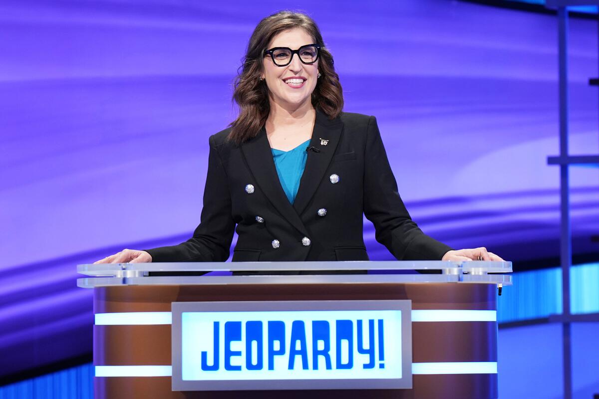 'Jeopardy' host Mayim Bialik wears a black blazer and black-rimmed glasses at the game show podium. 