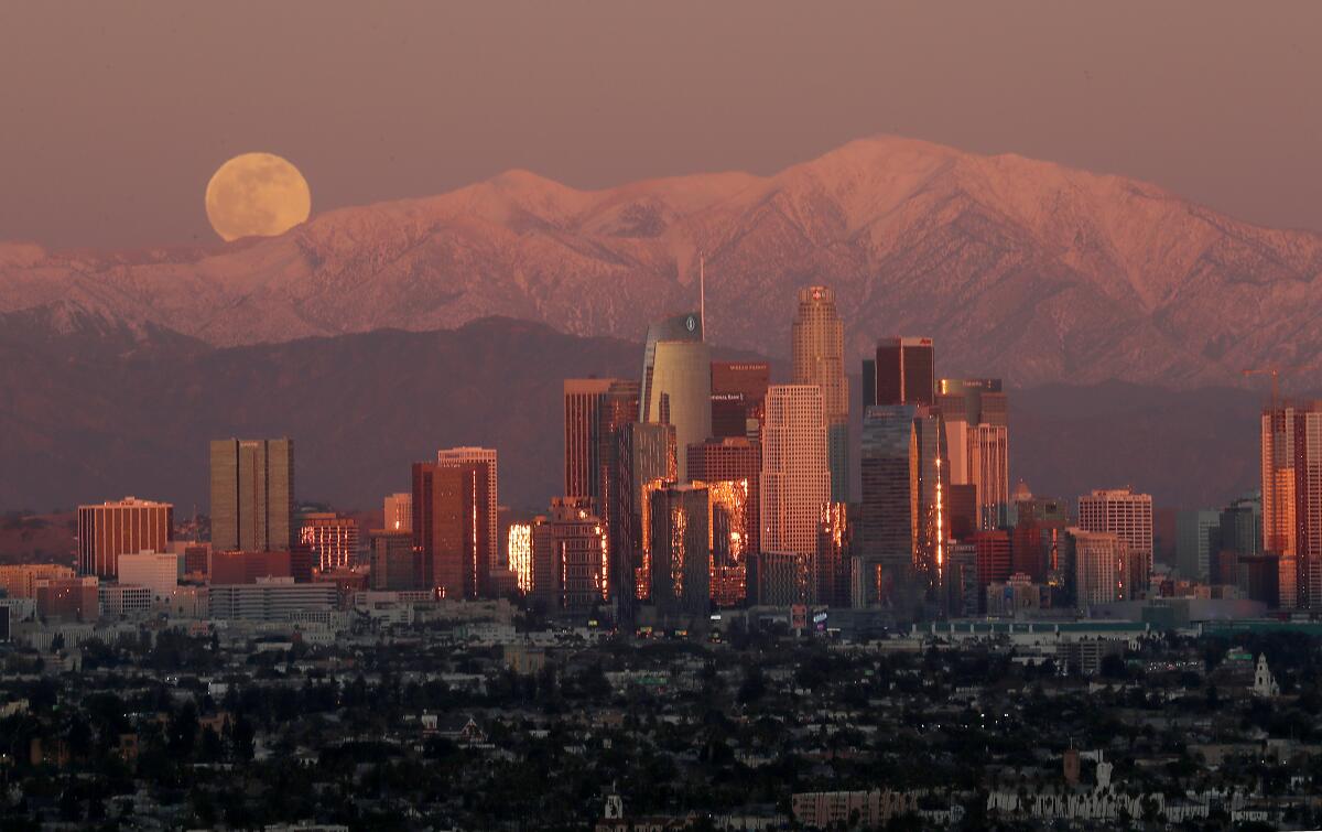 A full moon rises over the snow-capped San Gabriel Mountains and the skyline of downtown Los Angeles.