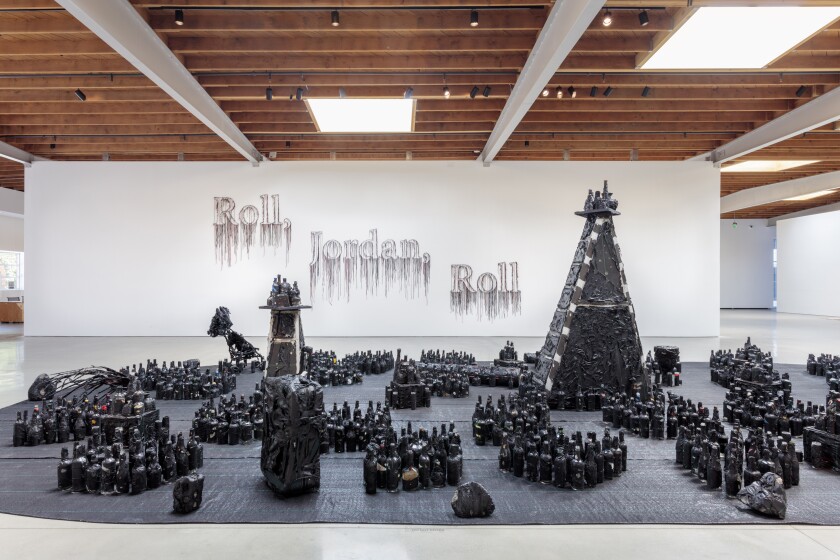 A black tarp on the floor of a gallery features arrangements of bottles and stepladders wrapped in a similar material.