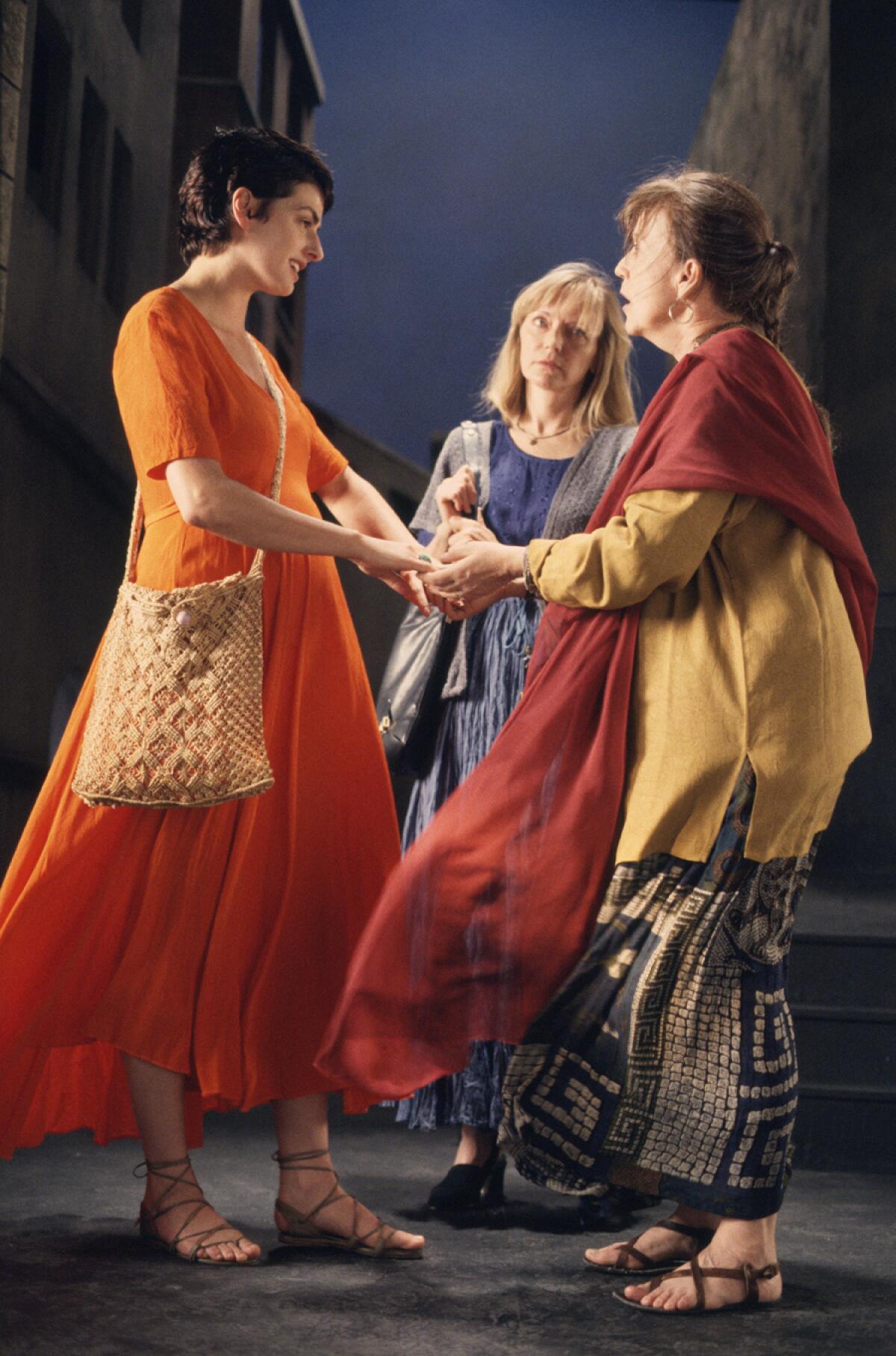 Dressed in flowing, brightly colored garments, two women warmly approach one another on the street as a third observes.