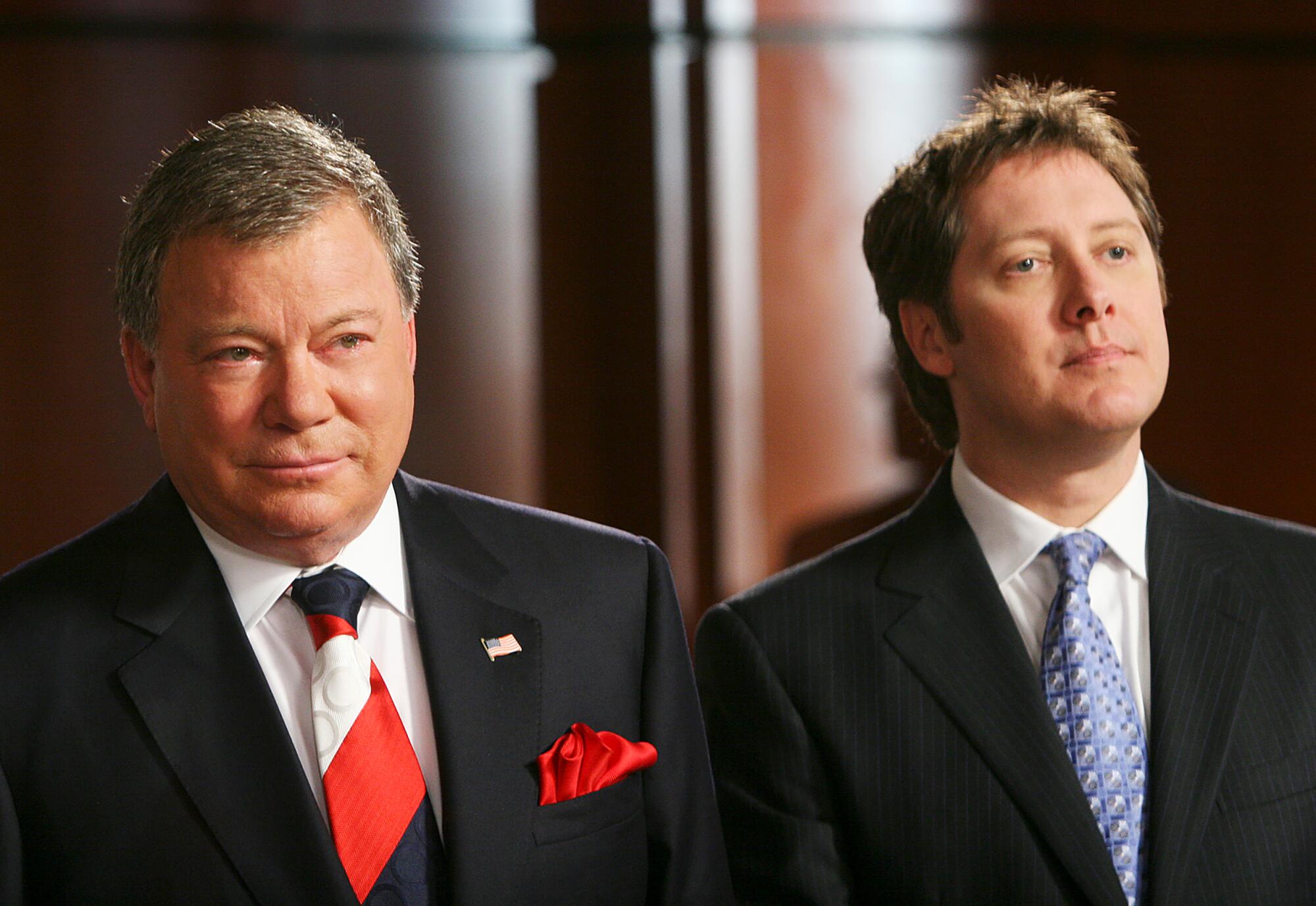 Two men stand side by side in black suits and ties.