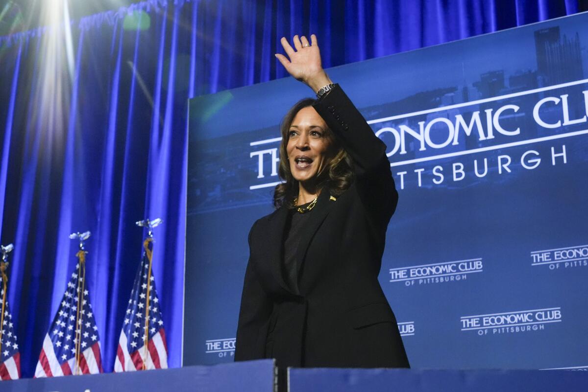 A woman with dark hair, wearing a dark suit, waves near U.S. flags, with a blue screen and blue curtains as a backdrop