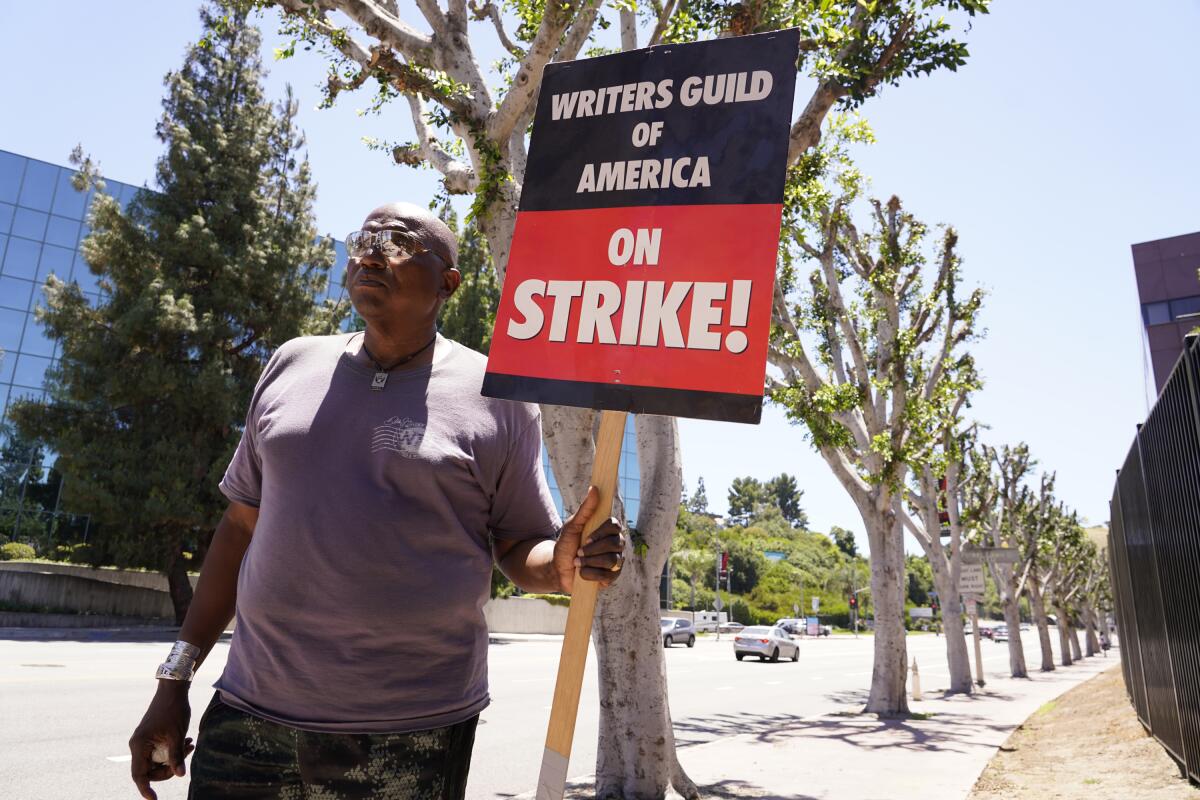 A picketer carries a sign that says Writers Guild of America on Strike!  near some trees. 