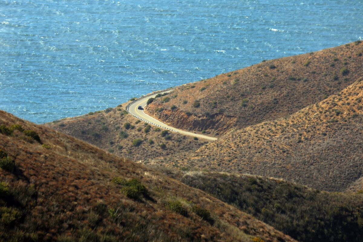 A road weaves through rolling green and brown hills by the ocean