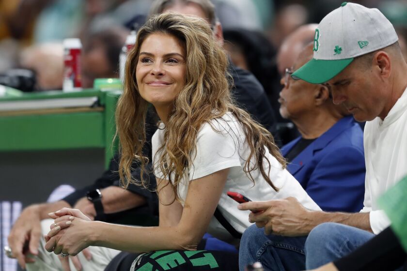 A smiling woman with long hair and a white T-shirt sits casually at a professional basketball game