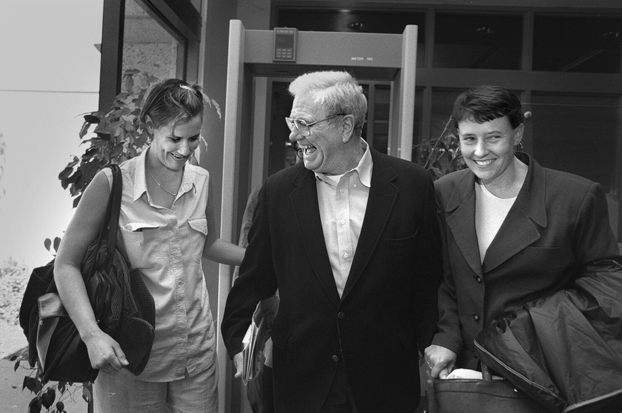 A black-and-white photo of George Franklin laughing, flanked by two smiling women as he walks past a security point