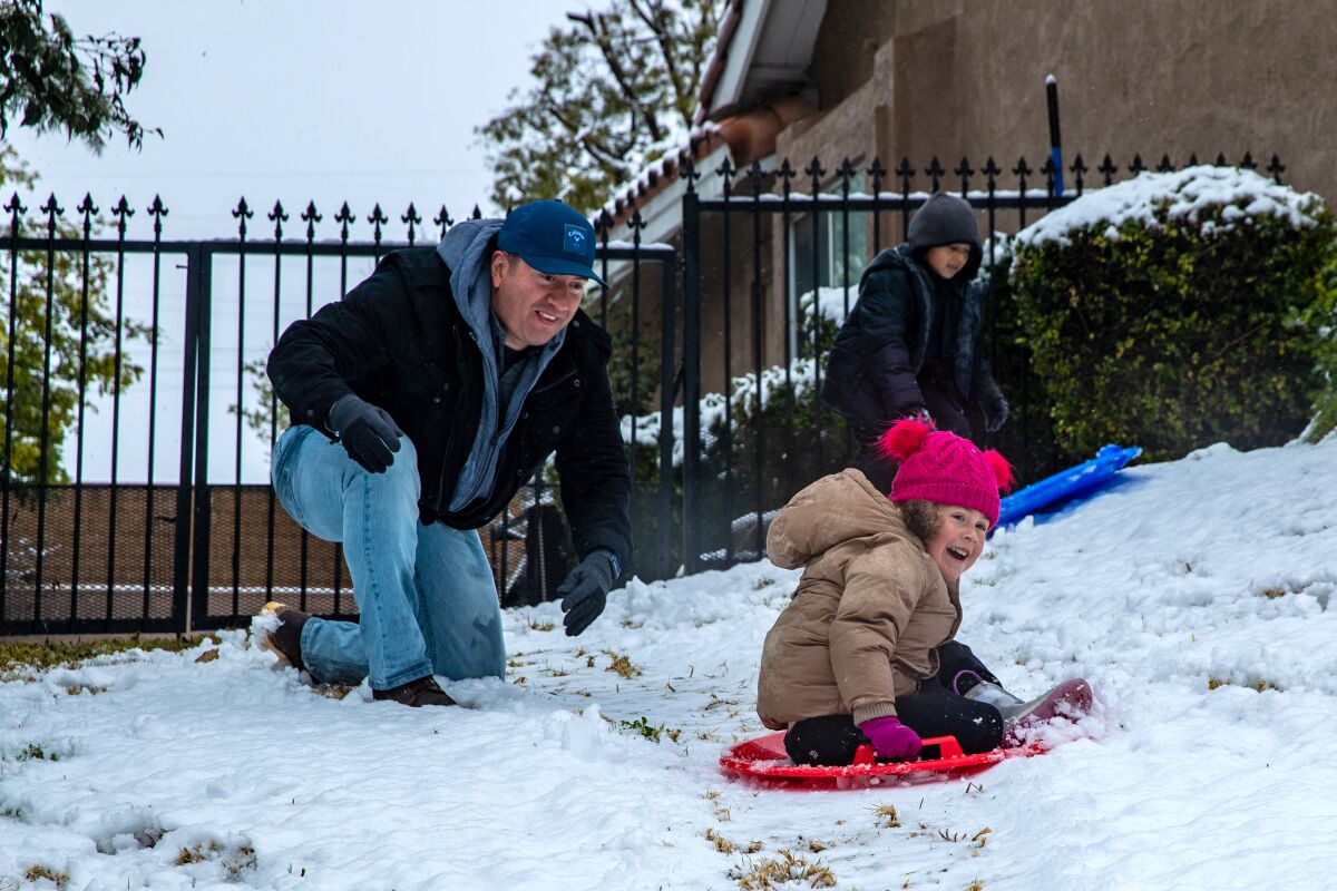 A man kneels as a child sleds down a small hill of snow.