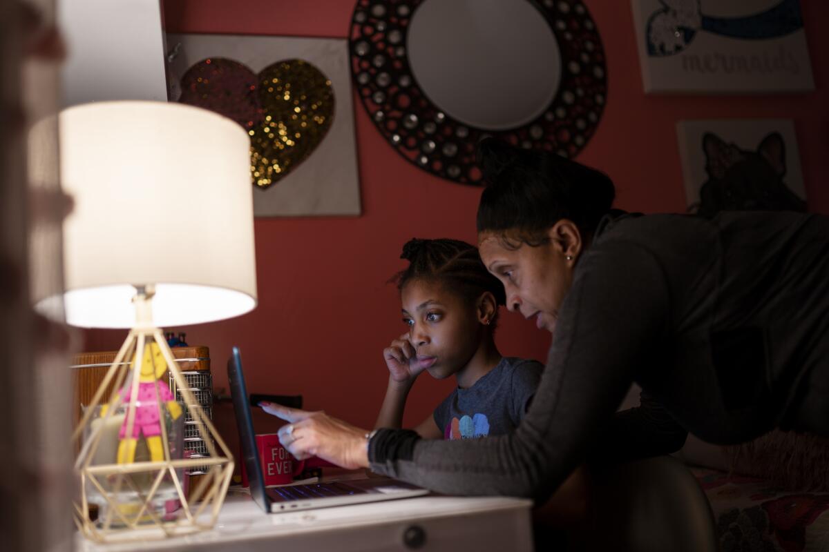 An 8-year-old girl, seated at a desk, stares attentively into her laptop screen as her mother looks at it too