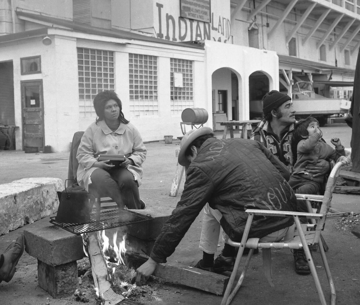 A woman, two men and a child sit in lawn chairs in front of a large white building and next to a small fire with a grate.