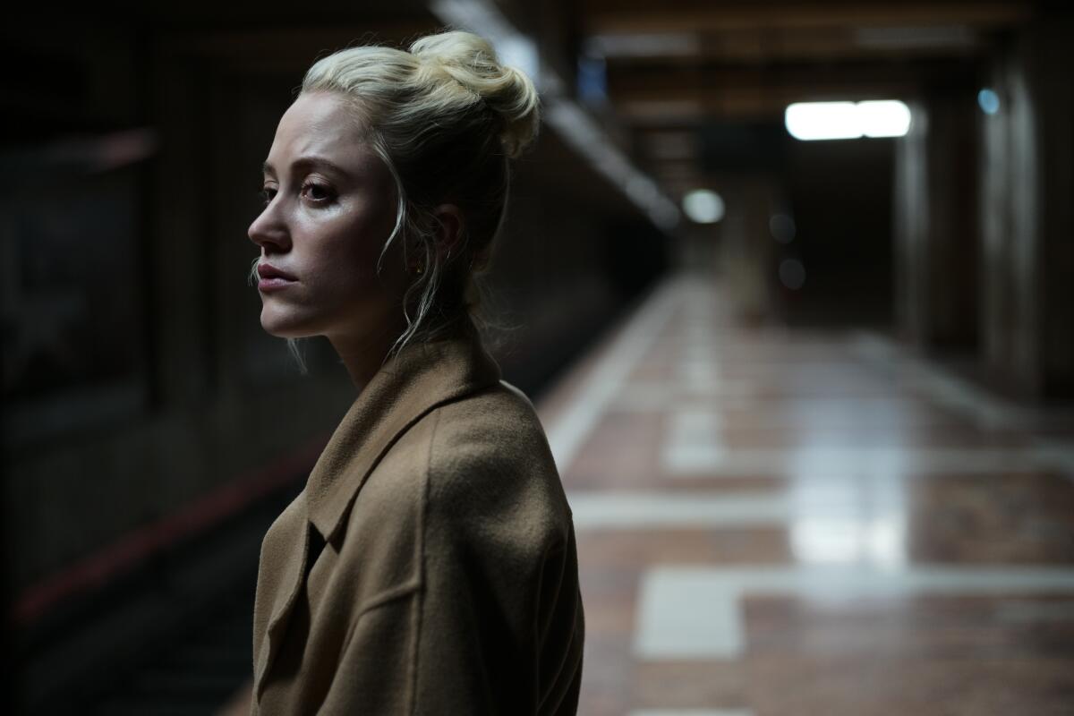 A blond woman with upswept hair and wearing a winter coat stands on an empty subway platform.