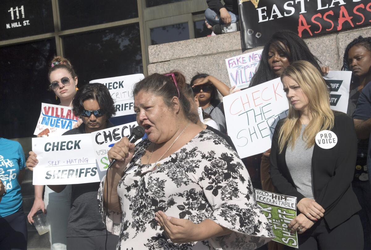 A woman speaks into a microphone. Behind her people hold signs including "Check the sheriff."