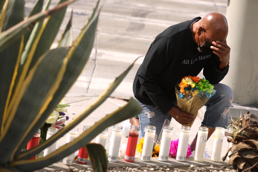 A man kneels and prays with flowers in one hand and his other hand over his eyes near a makeshift memorial on a sidewalk