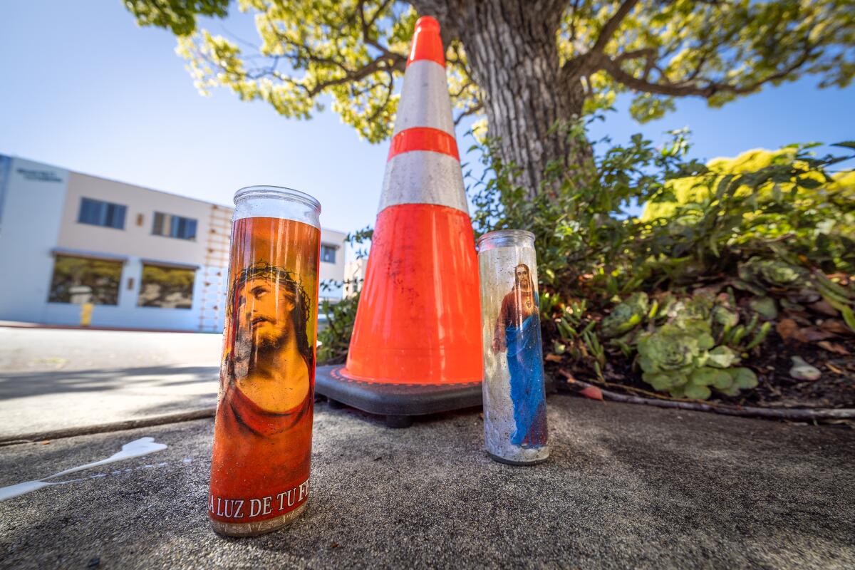 Devotional candles stand on a sidewalk beside an orange traffic cone 