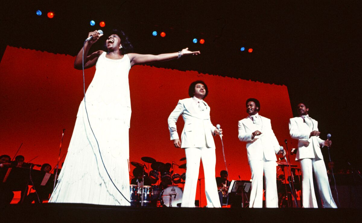 A singing group of one woman and three men, all dressed in white, performs onstage.
