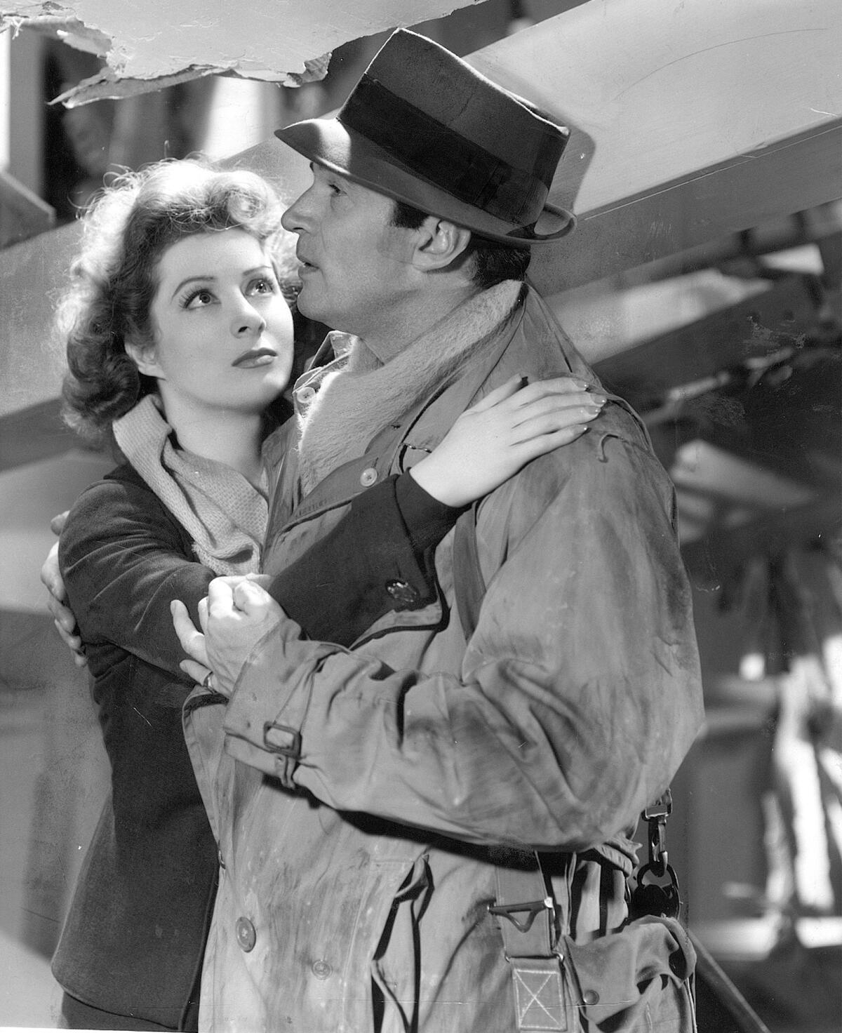 A black-and-white movie still of a woman with her arms around a man in a hat and trenchcoat, gazing up at him.